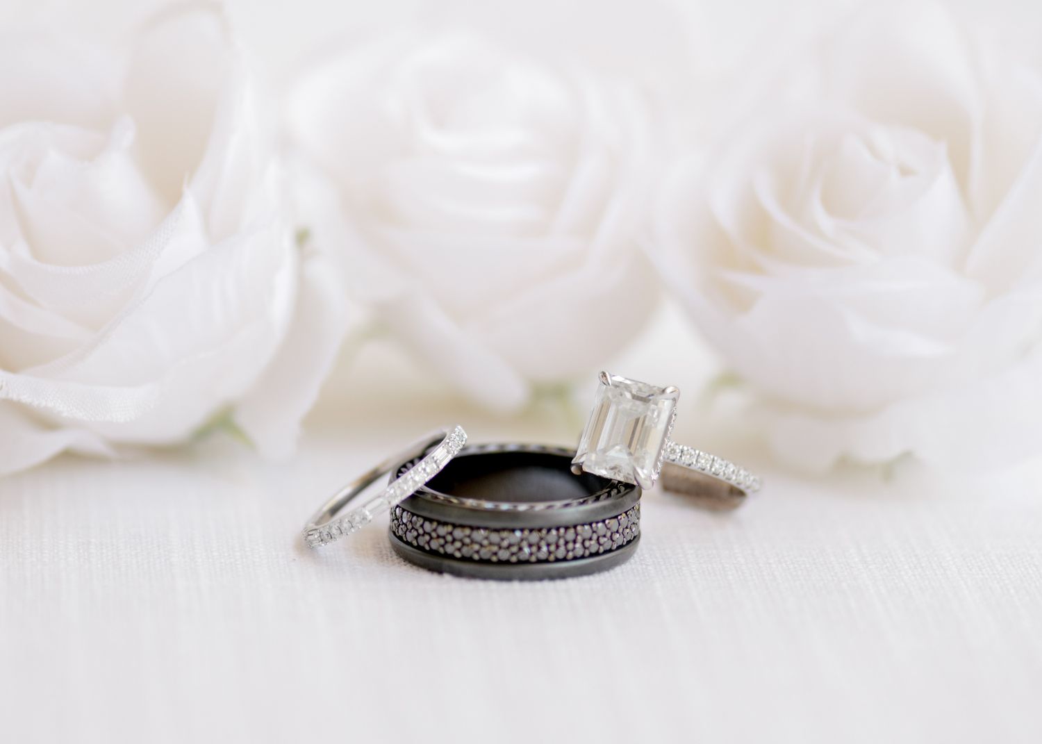 A delicate silver wedding band with pave diamonds rests against white rose petals in a soft romantic setting.