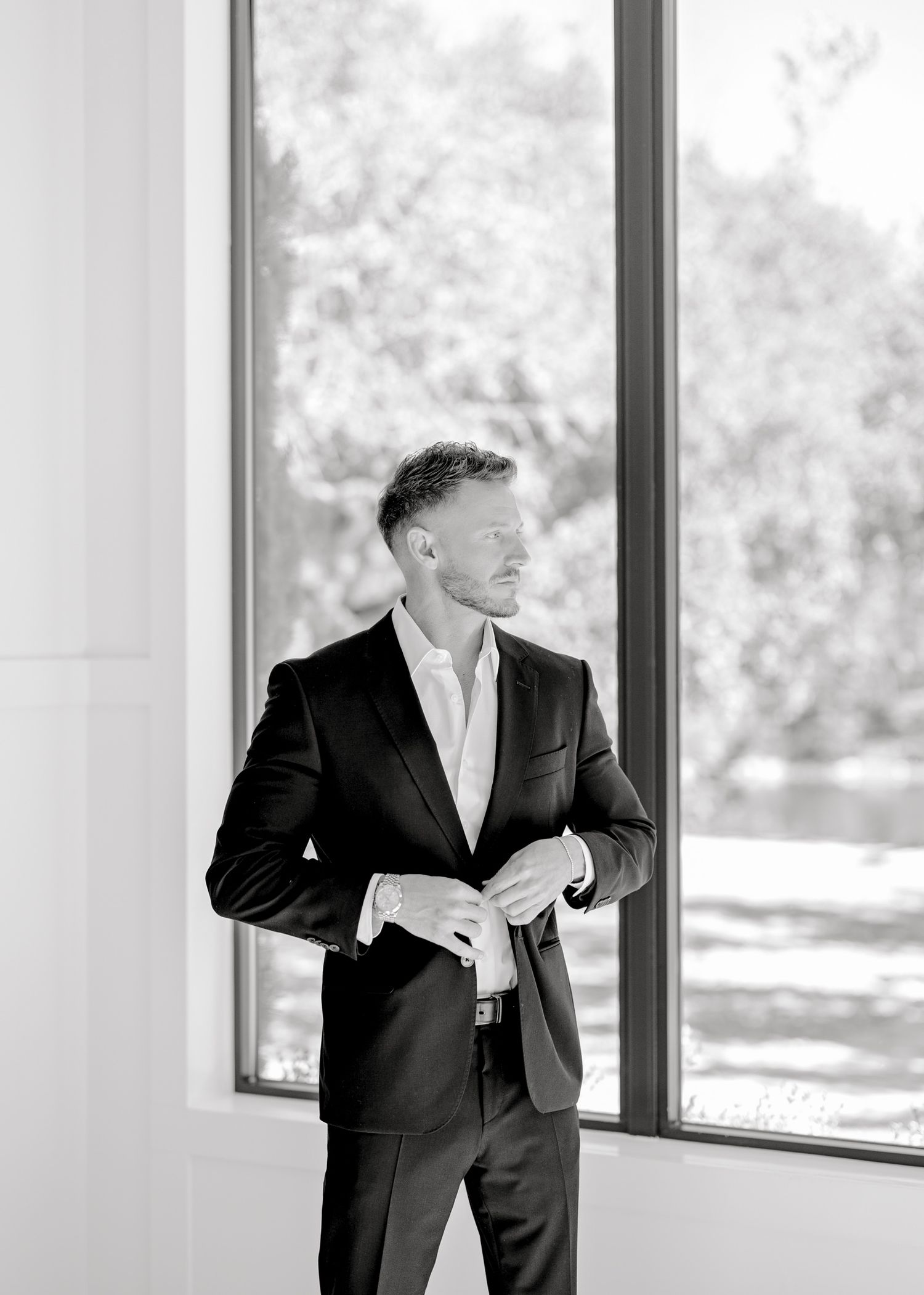 A man in a black suit and white shirt stands by large windows in a modern building interior.