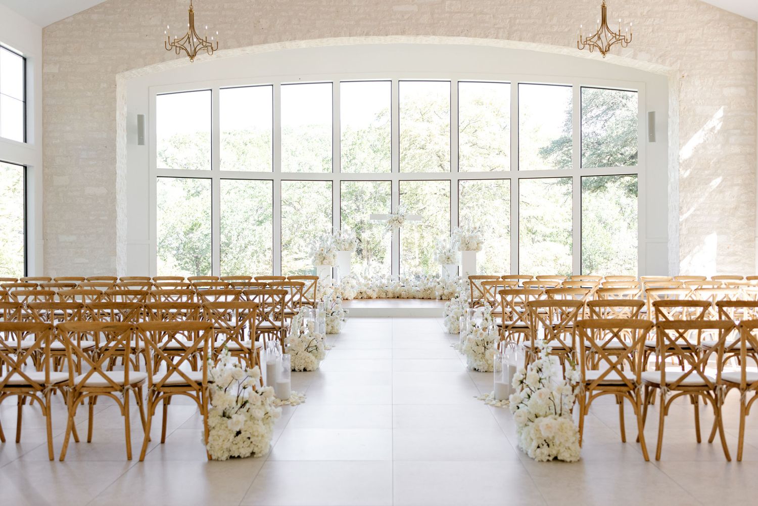 White wedding venue with wooden cross-back chairs arranged in rows and large windows letting in natural light.