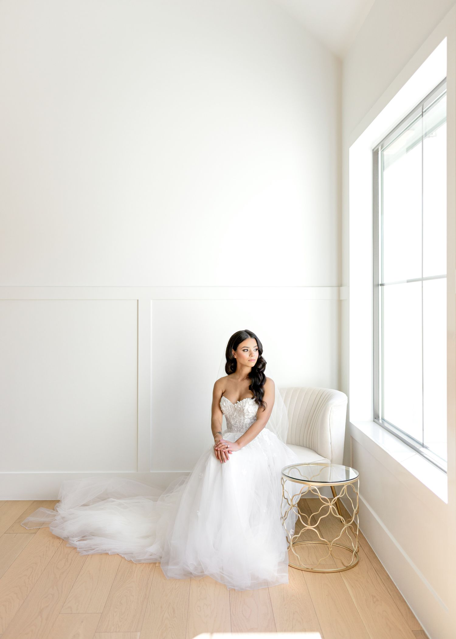 A bride in a flowing white gown poses elegantly in a bright minimalist studio with a gold accent table.
