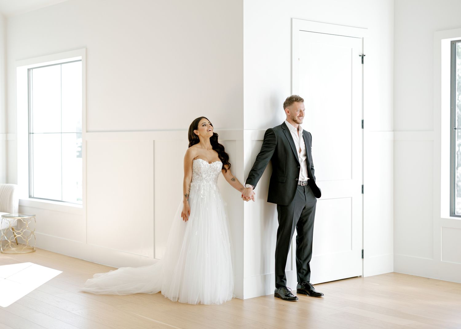 A couple holds hands while standing on opposite sides of a white door in a bright, minimalist room.