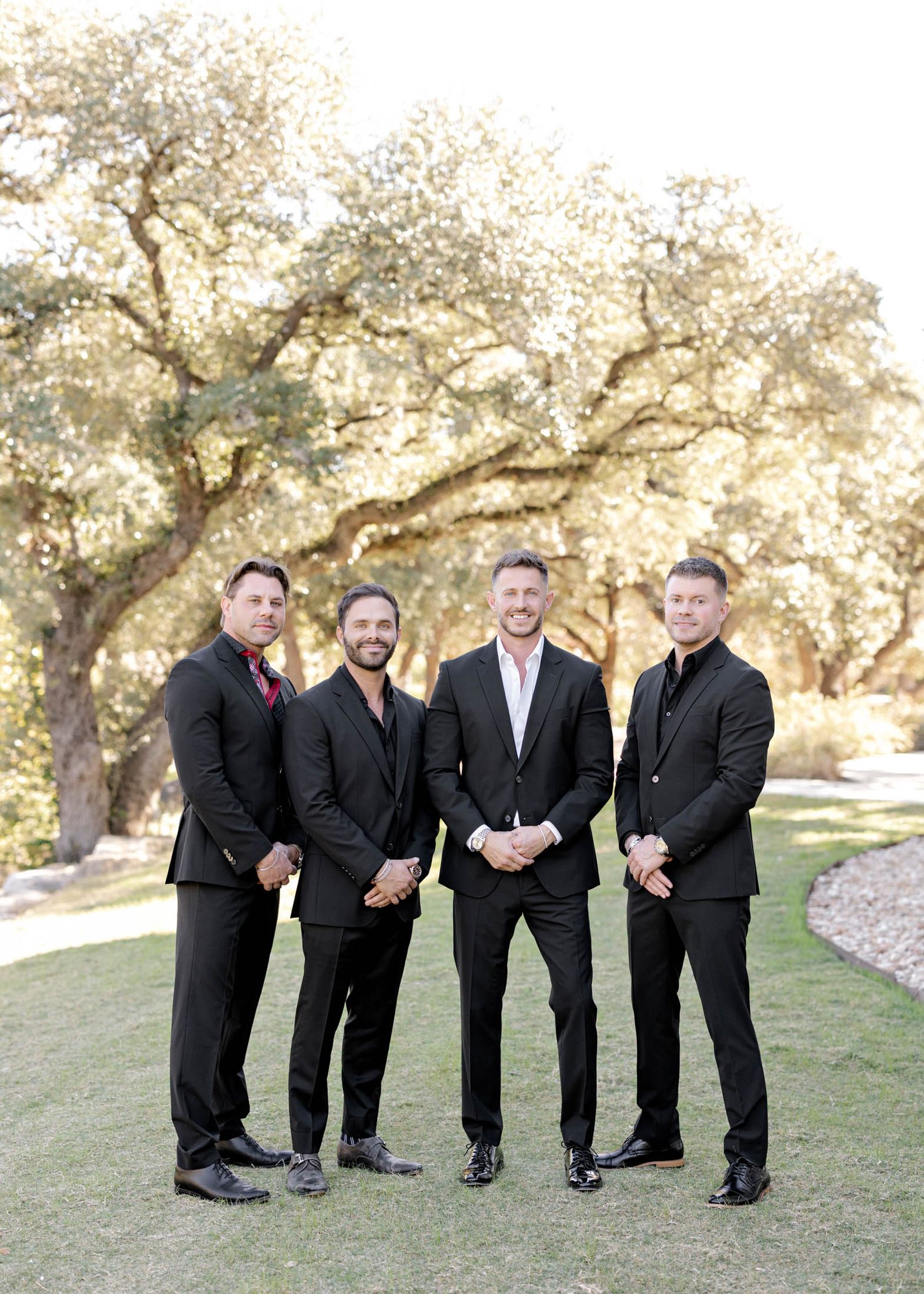 A group of groomsmen in black suits share a lighthearted moment while posing outdoors under oak trees.