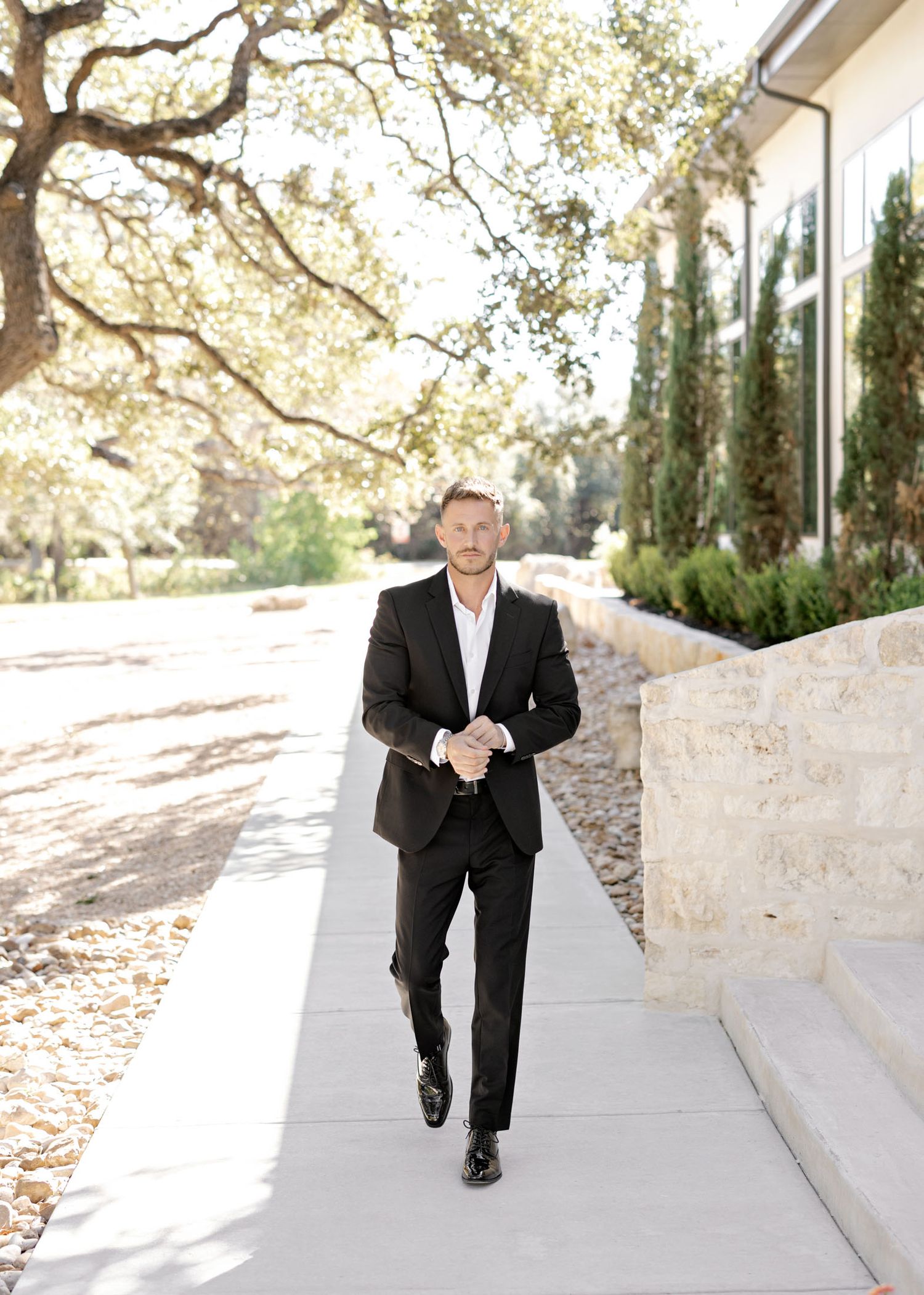 Person in black suit and white shirt walks confidently along a sunlit pathway with trees and architecture.