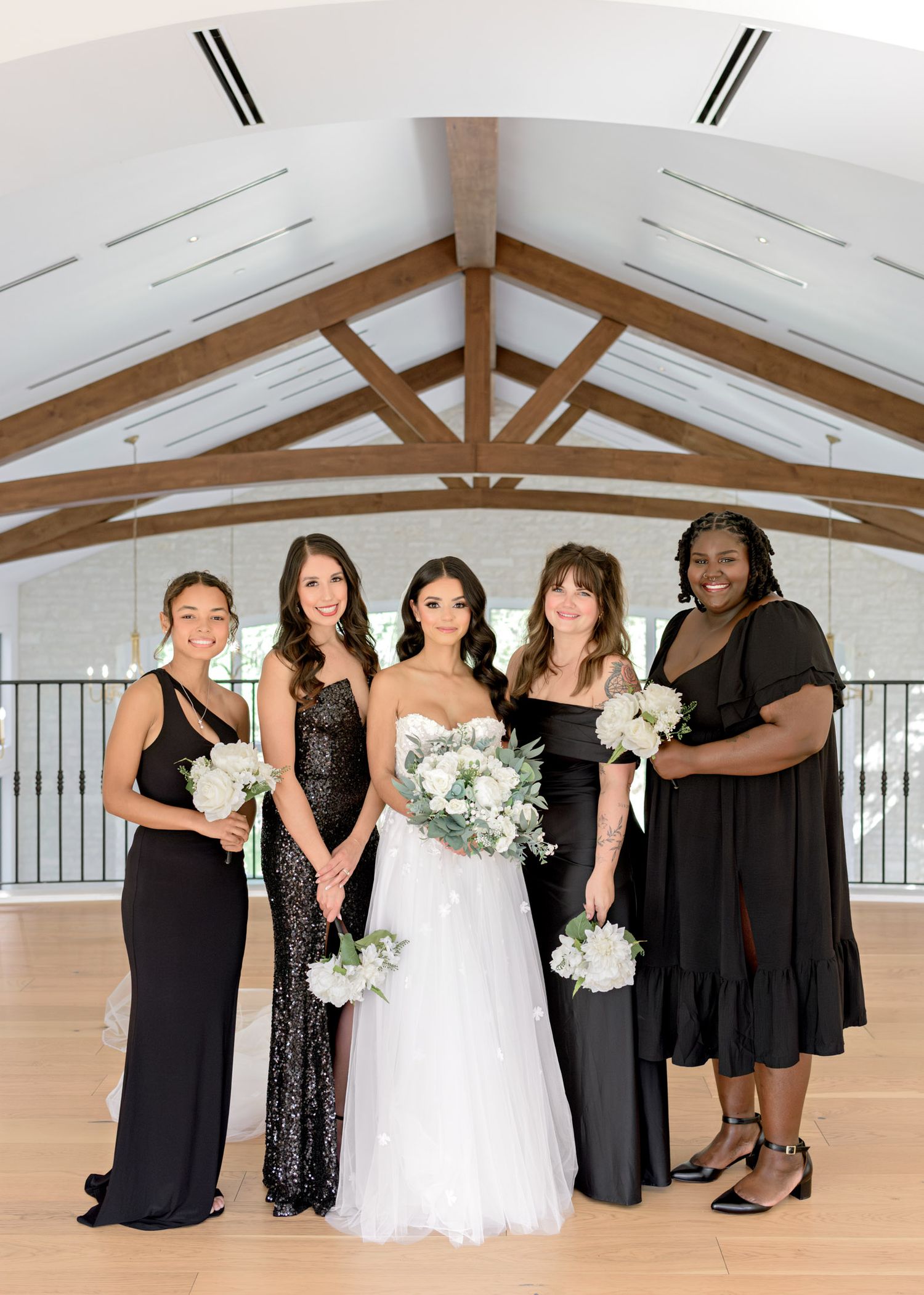 A bridal party in black dresses and a white wedding gown pose together in a bright indoor venue with wooden beams.