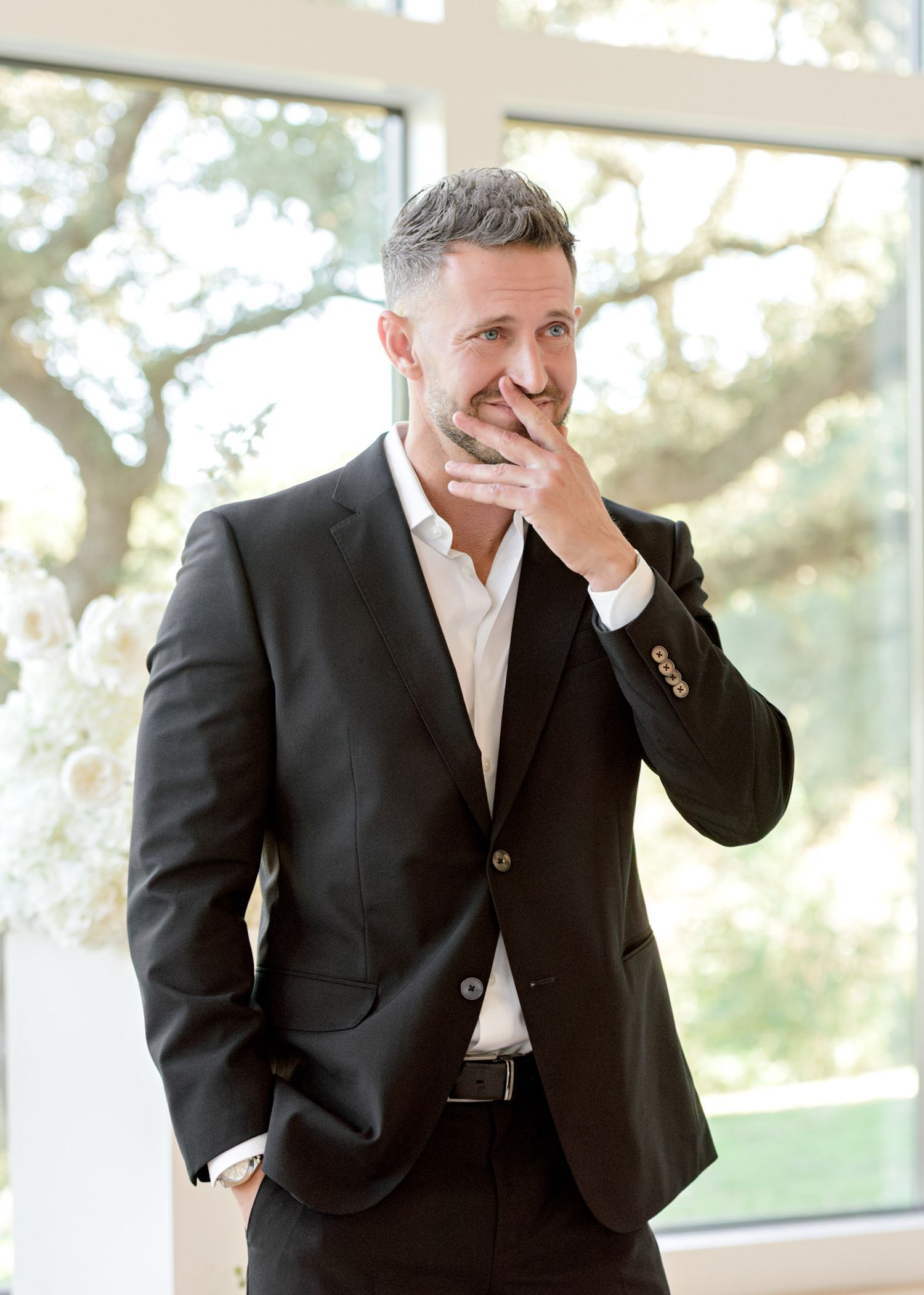 Professional in a dark suit laughs heartily while covering his face in a bright, window-lit setting.