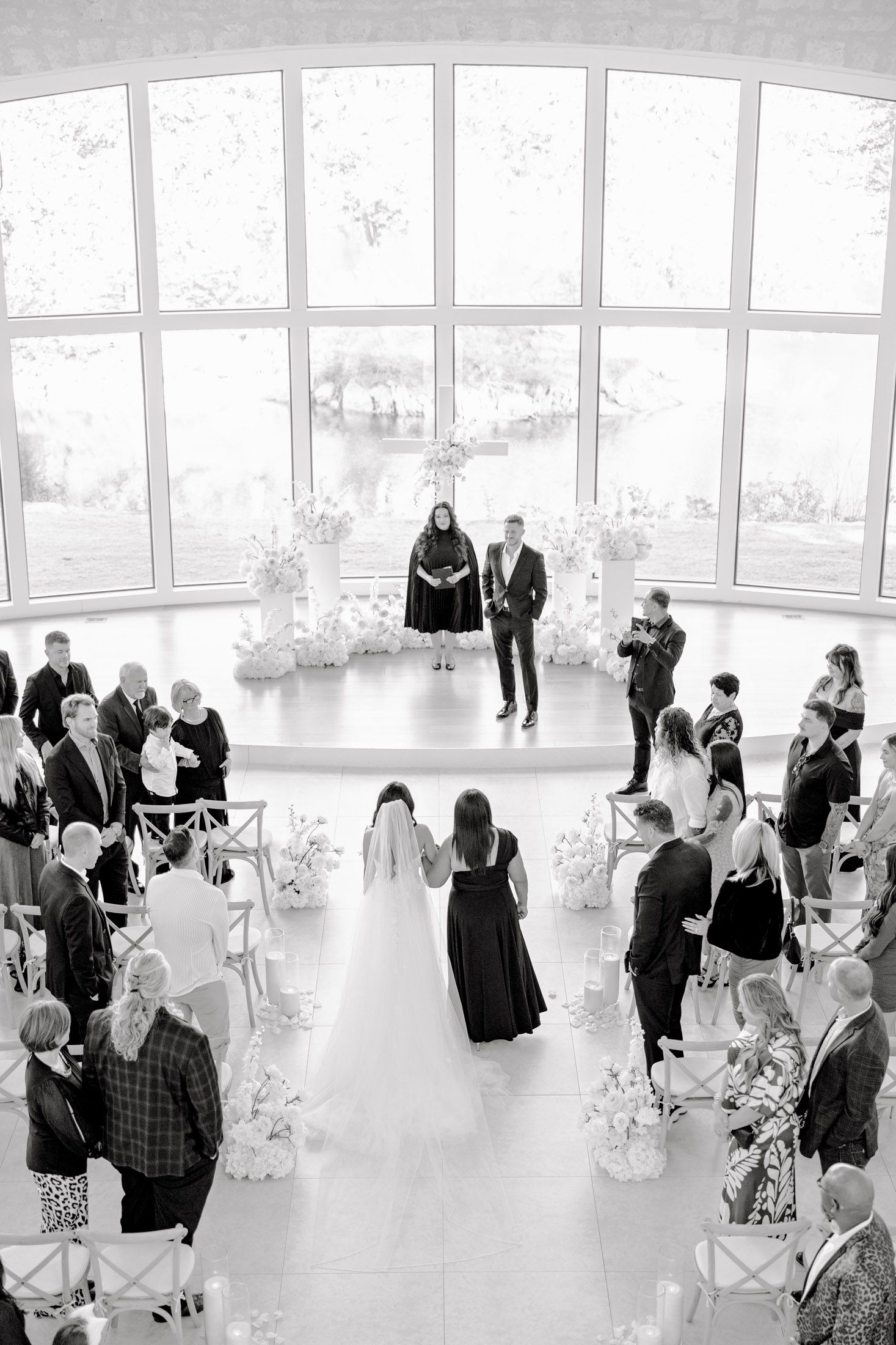 Black and white panoramic photo of guests mingling at an elegant event in a bright, window-lined venue.