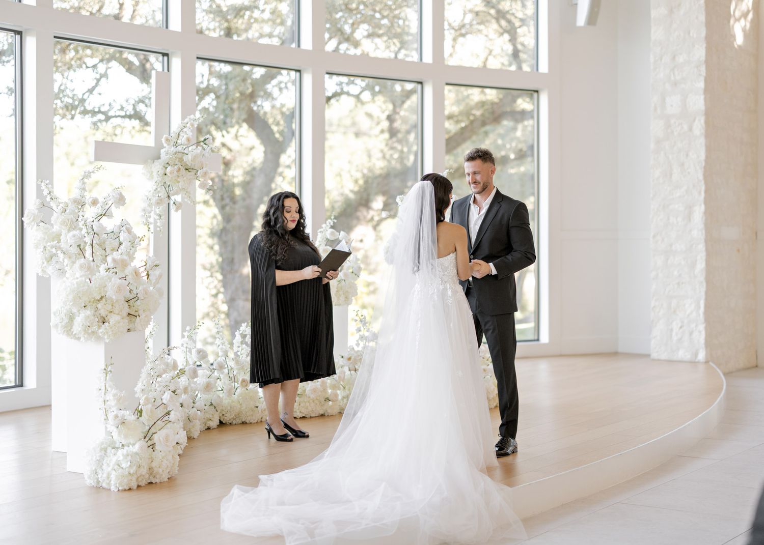Wedding ceremony in a bright, modern white room with floor-to-ceiling windows and floral decorations.