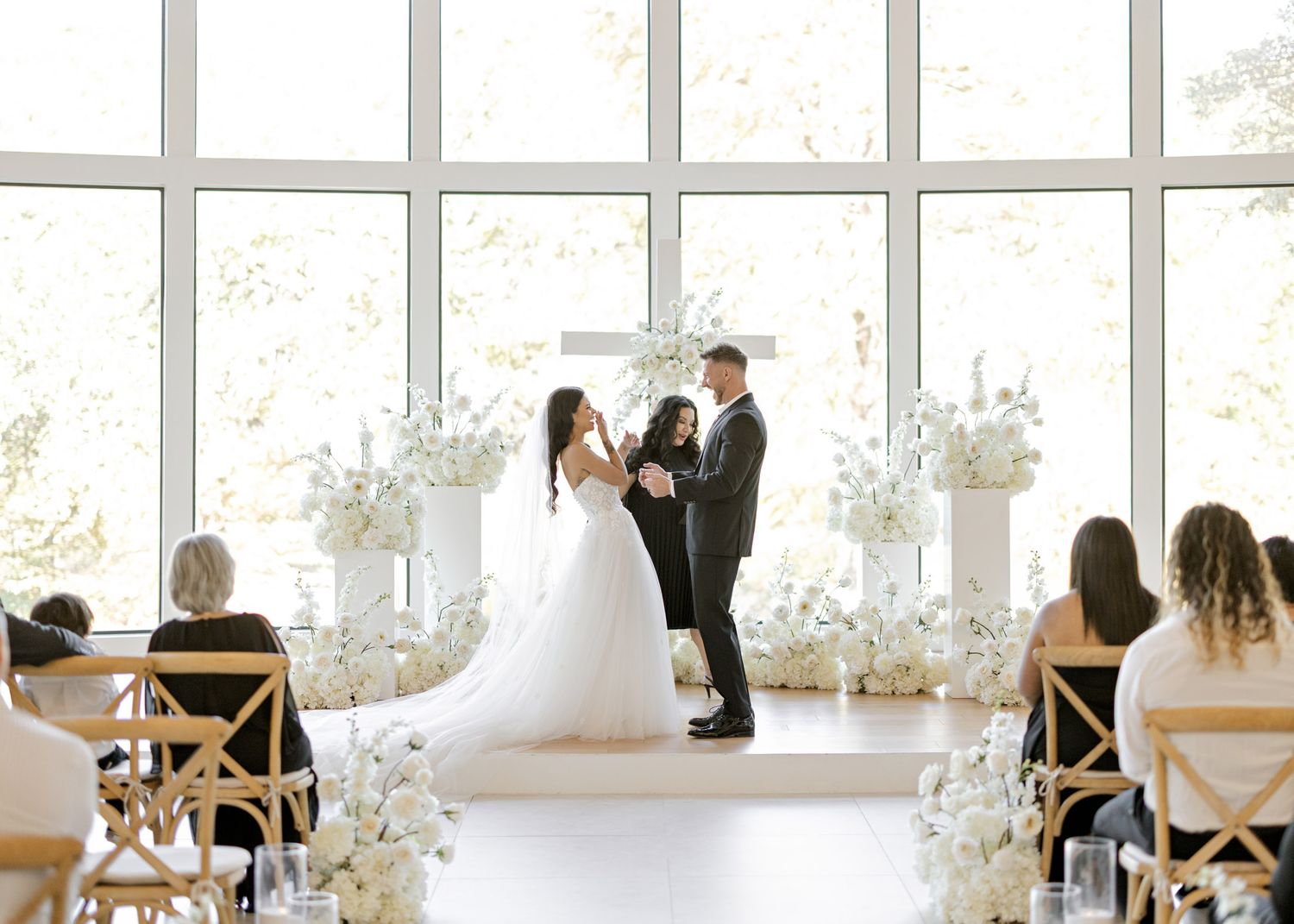 Wedding guests seated in wooden chairs watching ceremony in a sunlit room with white floral arrangements.