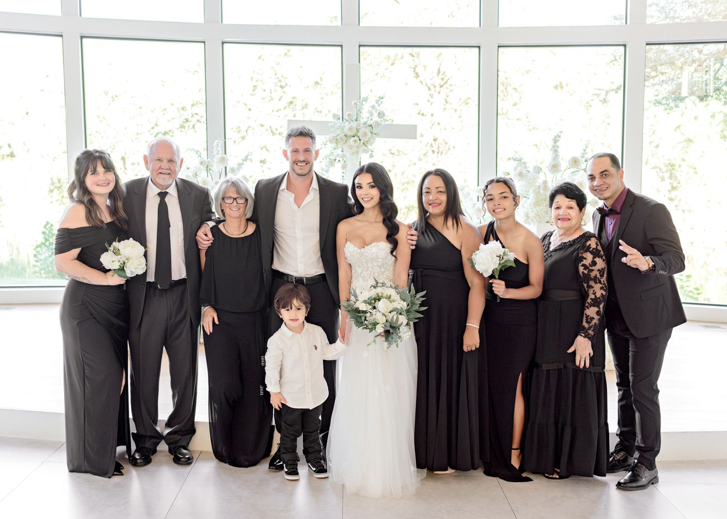 Wedding party in formal black and white attire poses together in bright indoor venue with large windows.