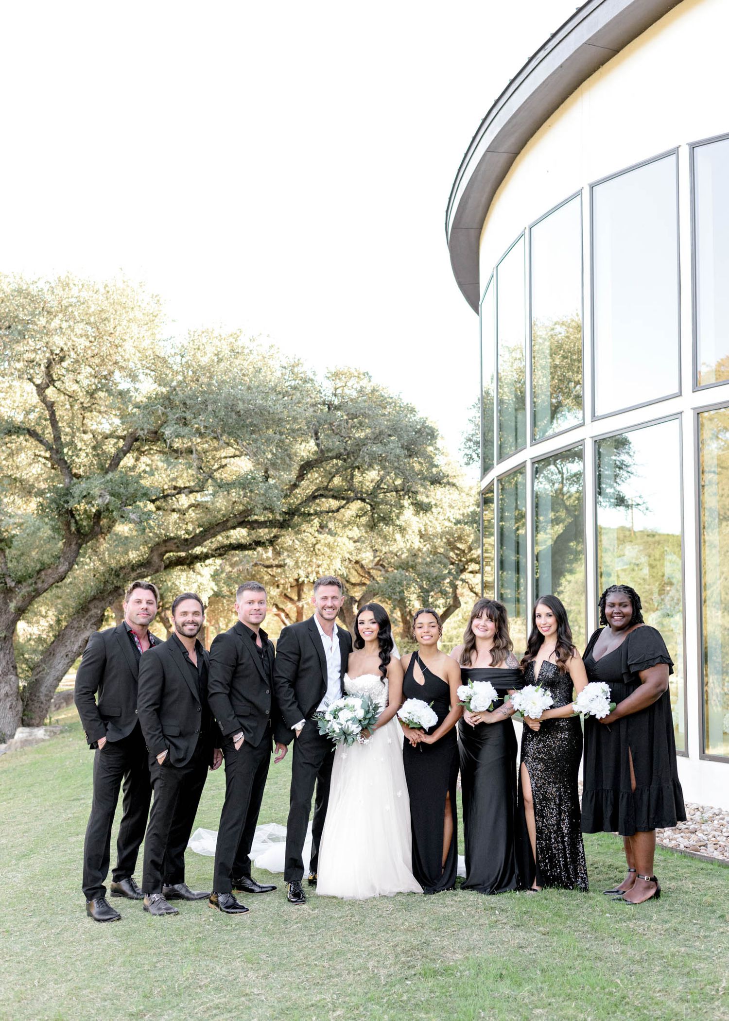 Wedding party in black attire poses together outside a modern glass building with trees in the background.