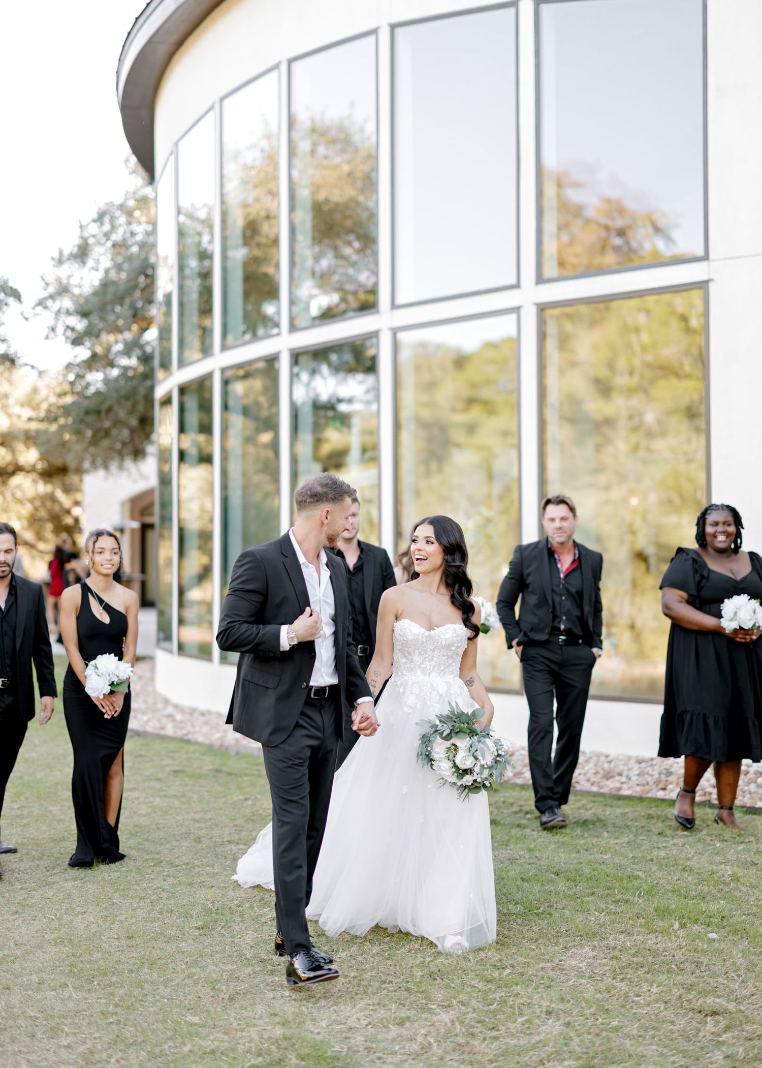 Newlyweds walk together outside a modern building with curved glass windows while wedding party follows behind.