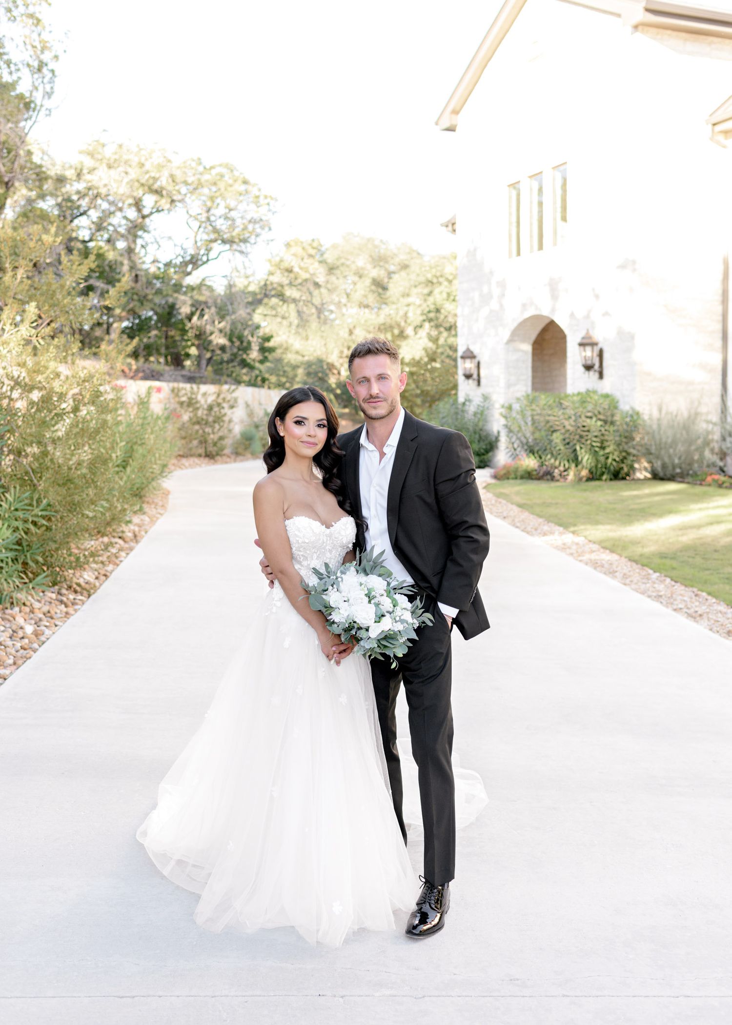 A couple in formal wedding attire poses together on a bright white walkway outside a building.