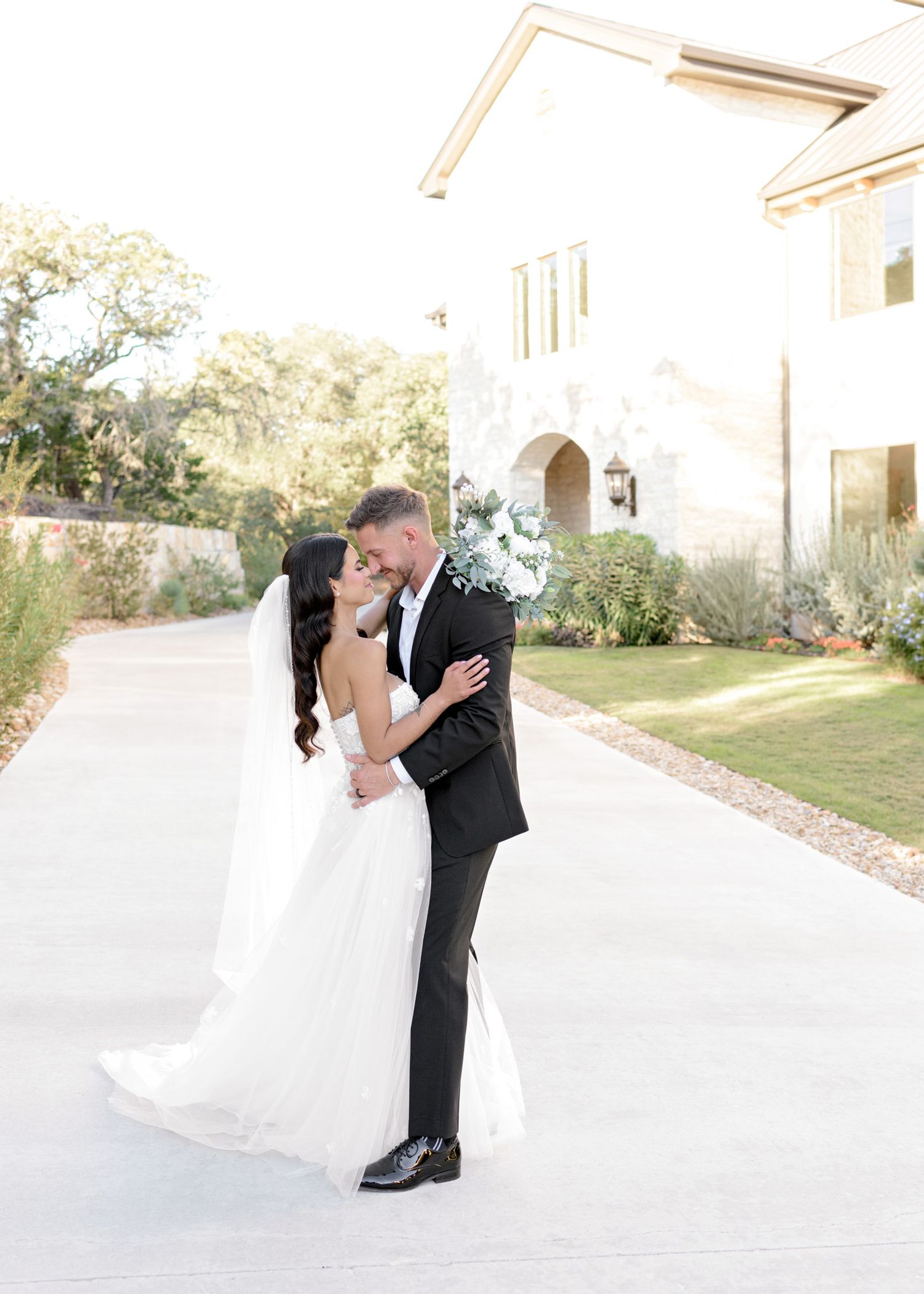 Romantic wedding moment captured on a sunlit driveway with white stucco building in background.