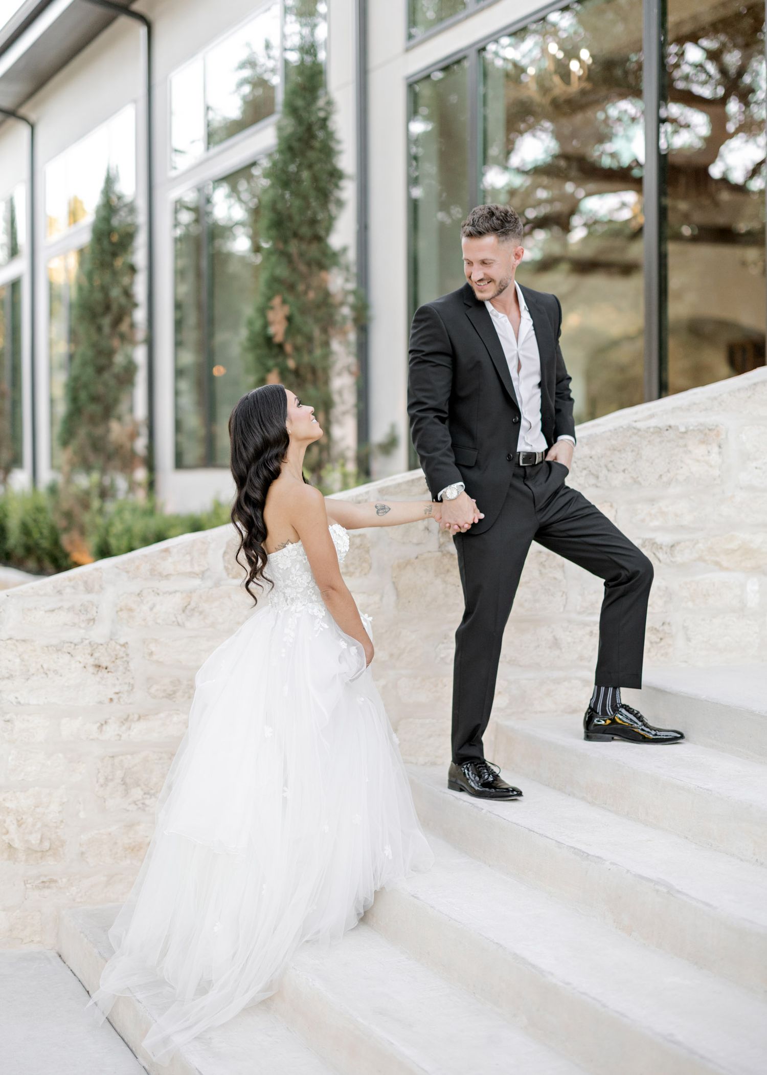 A bride in a white ballgown and groom in black suit pose on elegant stone steps outside a modern glass building.