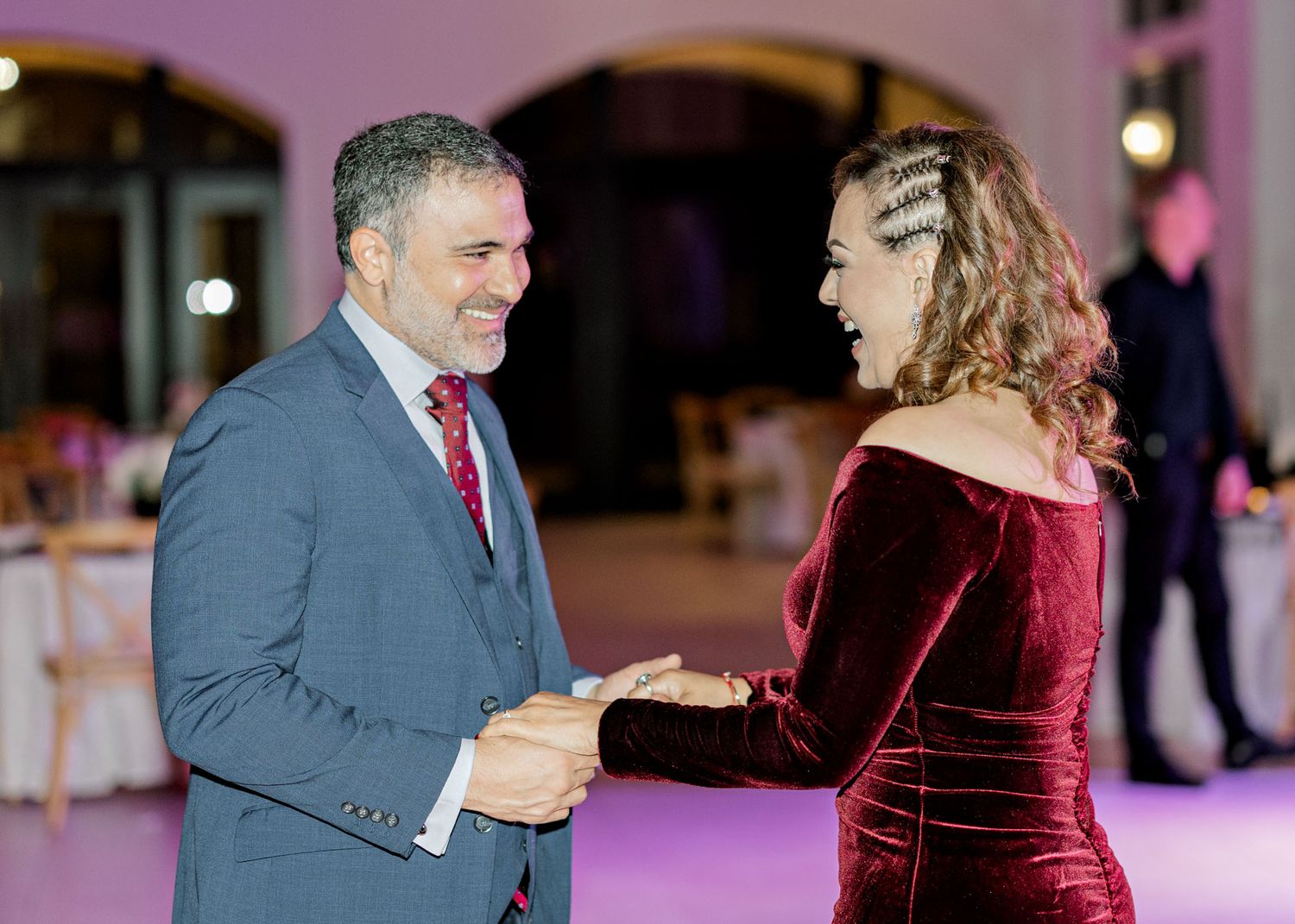 A couple in formal attire dances together at an event with pink lighting in the background.
