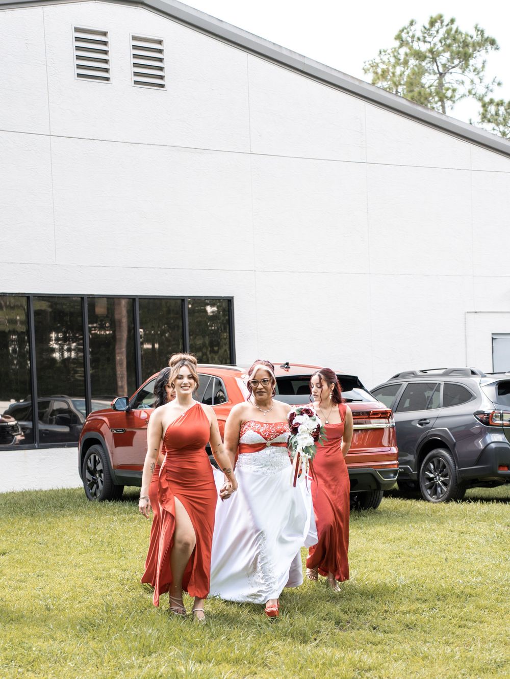 A bridal party walks across grass wearing coral-colored dresses outside a white building in Tampa Florida