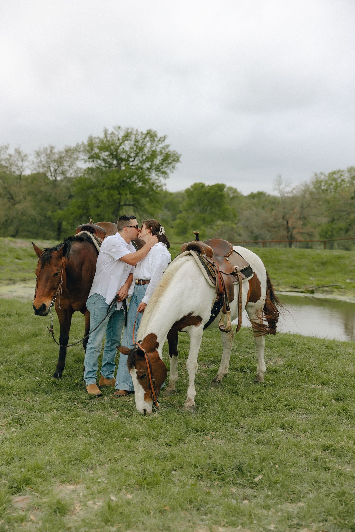Two people tend to horses on a grassy field near a pond on an overcast day.