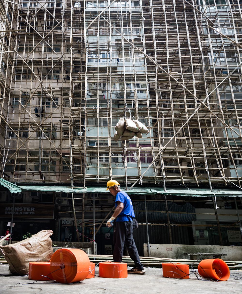 Construction worker in yellow hard hat walking past bamboo scaffolding on Hong Kong building site.
