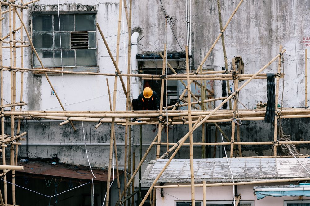 Construction worker in yellow helmet standing on bamboo scaffolding against weathered concrete building wall.