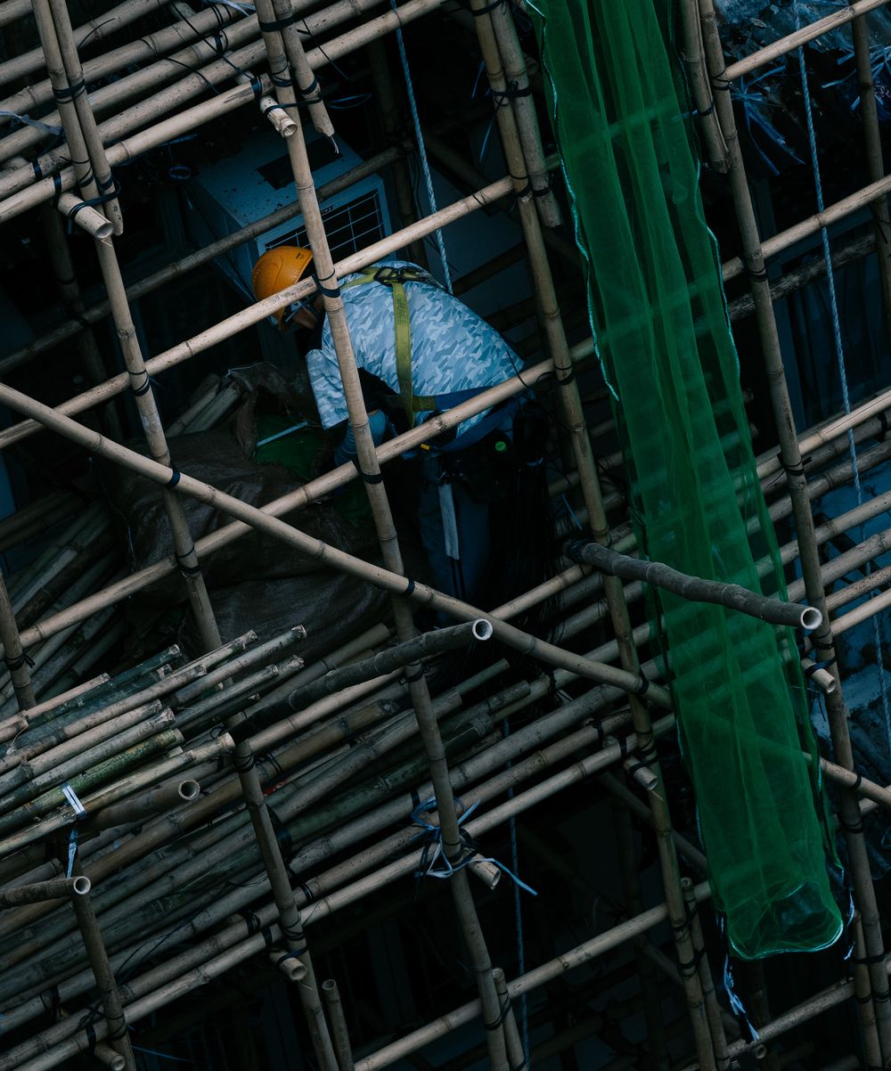 Worker in blue shirt and yellow helmet working within dense bamboo scaffolding on building exterior.
