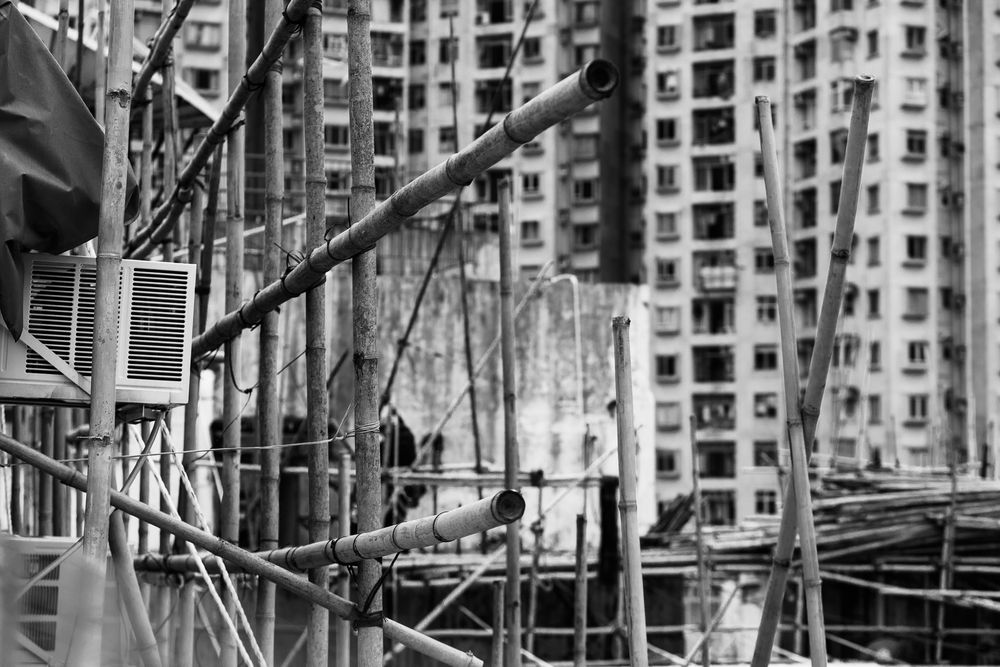 Black and white image of bamboo scaffolding with dense high-rise residential towers in the background.
