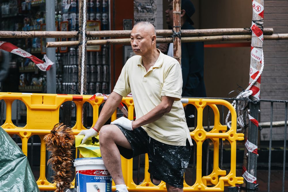 Middle-aged Asian man resting on a bucket beside yellow construction barriers on a city street.
