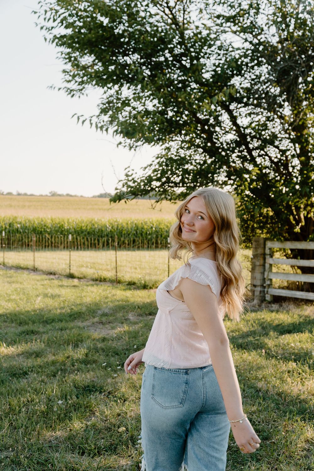 A person in light pink top and jeans stands in a grassy field at sunset with wooden fencing and cornfields in background.