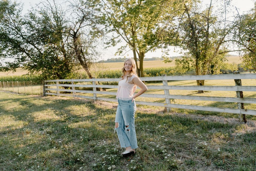 A person in a flowy dress walks through a golden sunlit meadow with white wooden fences in the background.