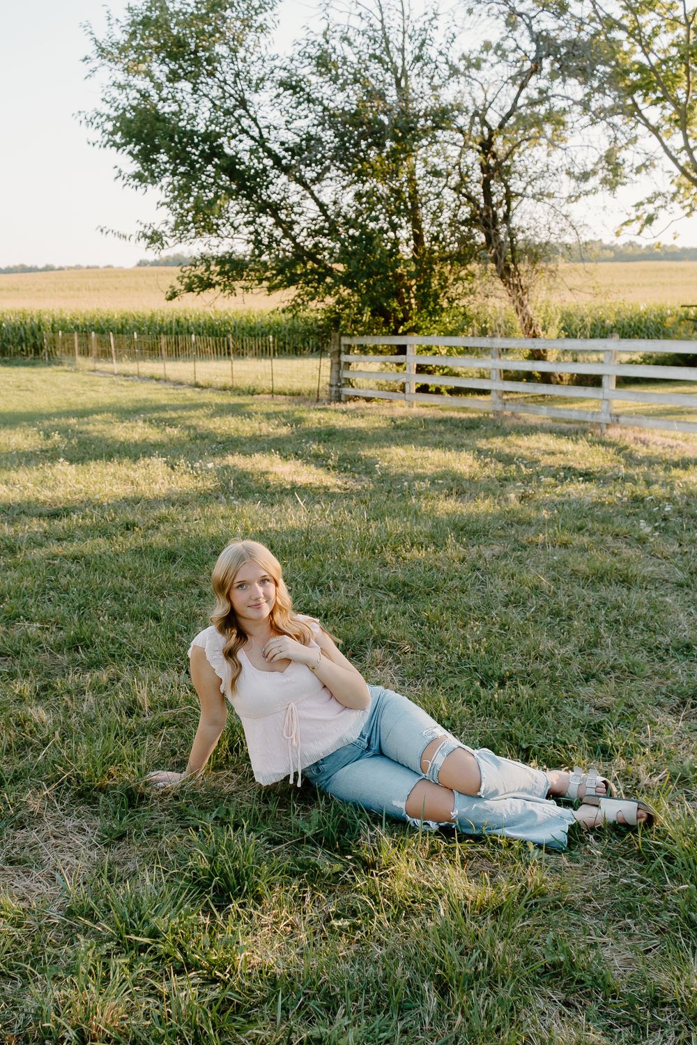A person relaxes in a grassy field on a sunny day with white fencing and trees in the background.
