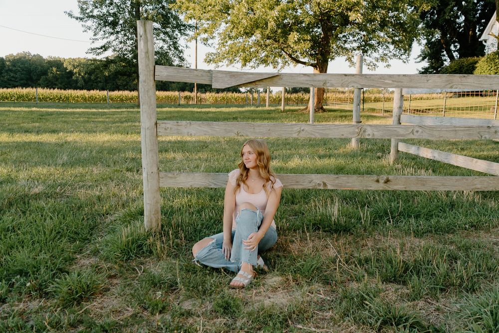 A person sits on a wooden fence post in a rustic country field at sunset.