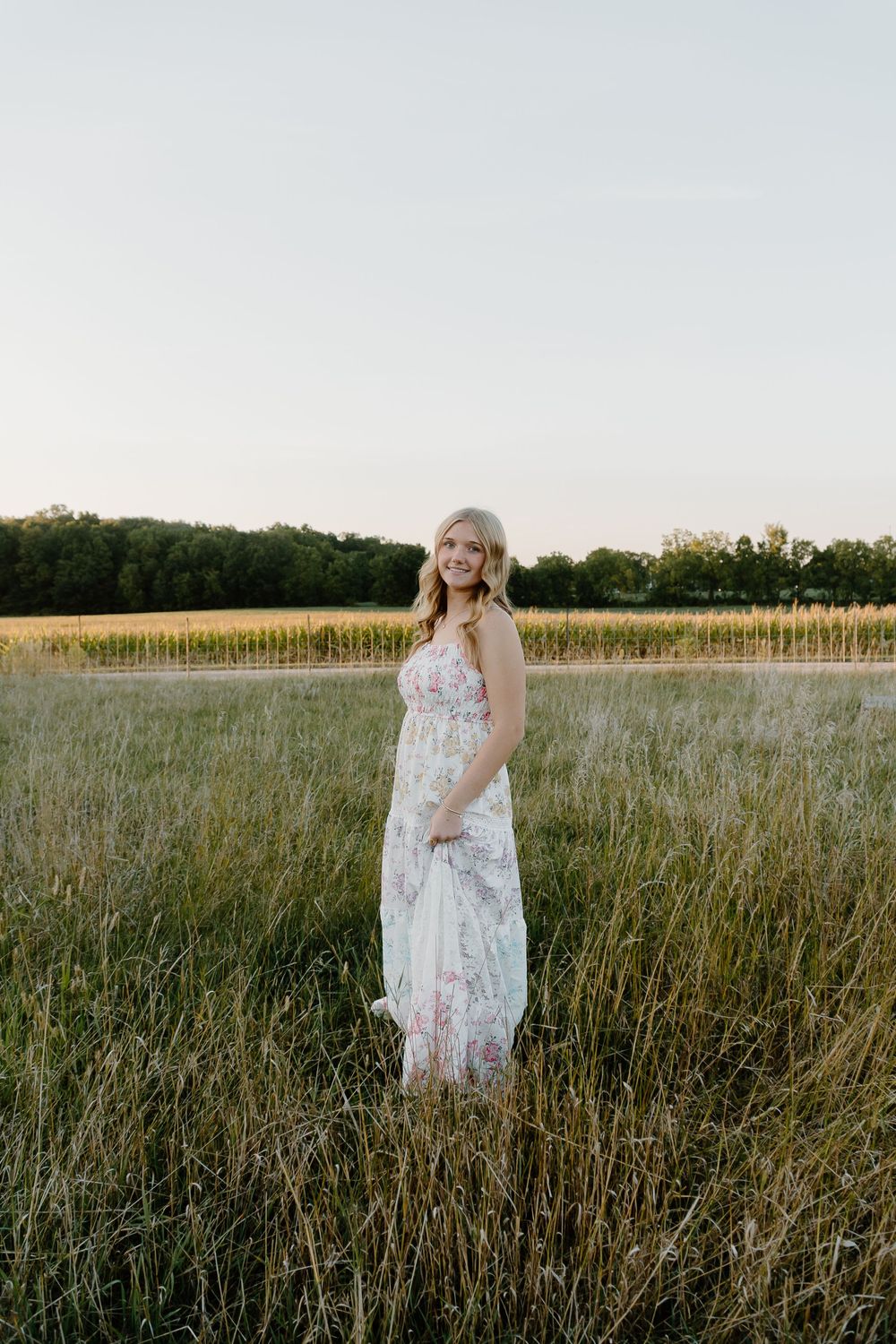A person in a white floral dress stands in a golden field at sunset with trees in the background.