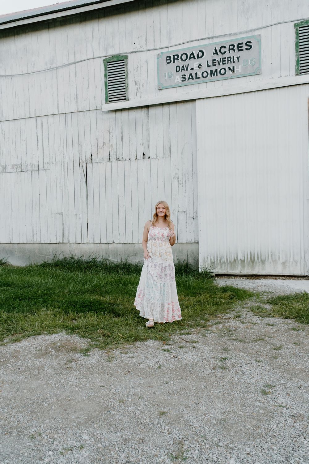 A person in a light-colored dress stands against a rustic white barn wall at Broad Acres.