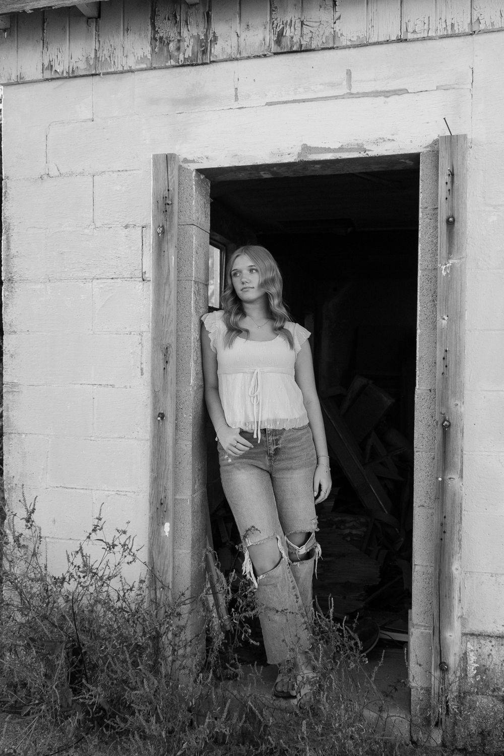 Black and white photograph of someone standing in a doorway of an old building with distressed walls and overgrown weeds.