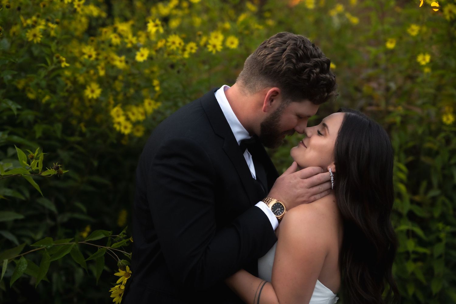 A joyful wedding couple shares a romantic kiss amid lush greenery and sunlit foliage in an outdoor garden setting.