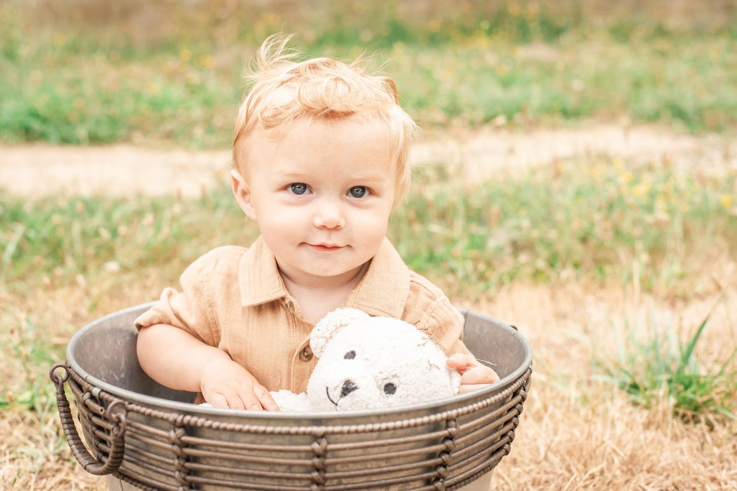 Small child sits in a round woven basket with a stuffed teddy bear toy on a sunny day in a grassy field.