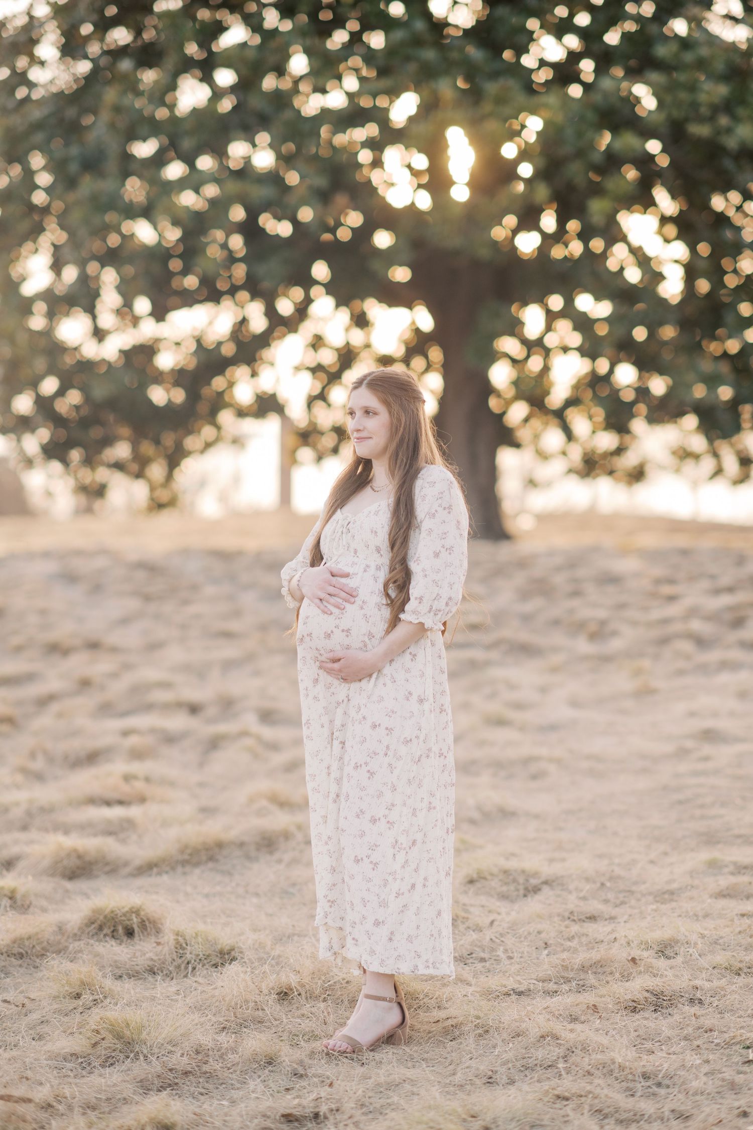 Pregnant woman in floral dress cradling her baby bump during golden hour maternity session.