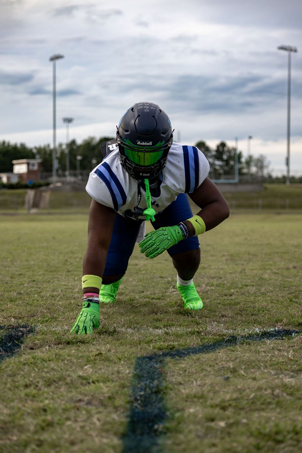 Football player wearing bright green gloves crouches in three point stance position on grass field.