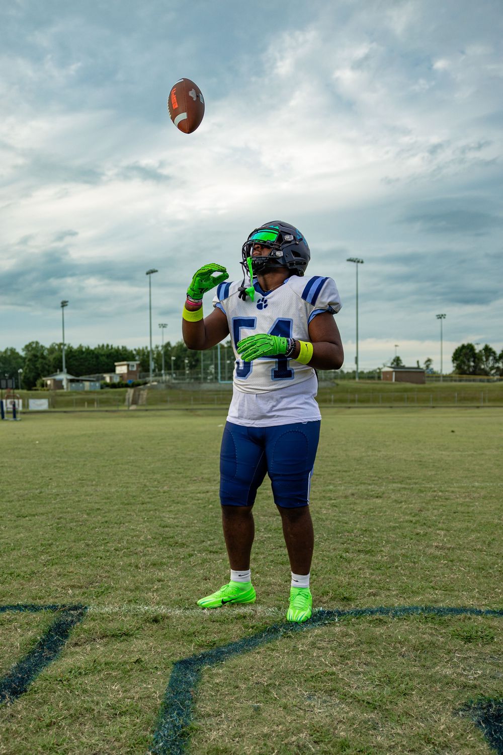 Football player practices catching drills with neon green gloves on a grass field under cloudy skies.