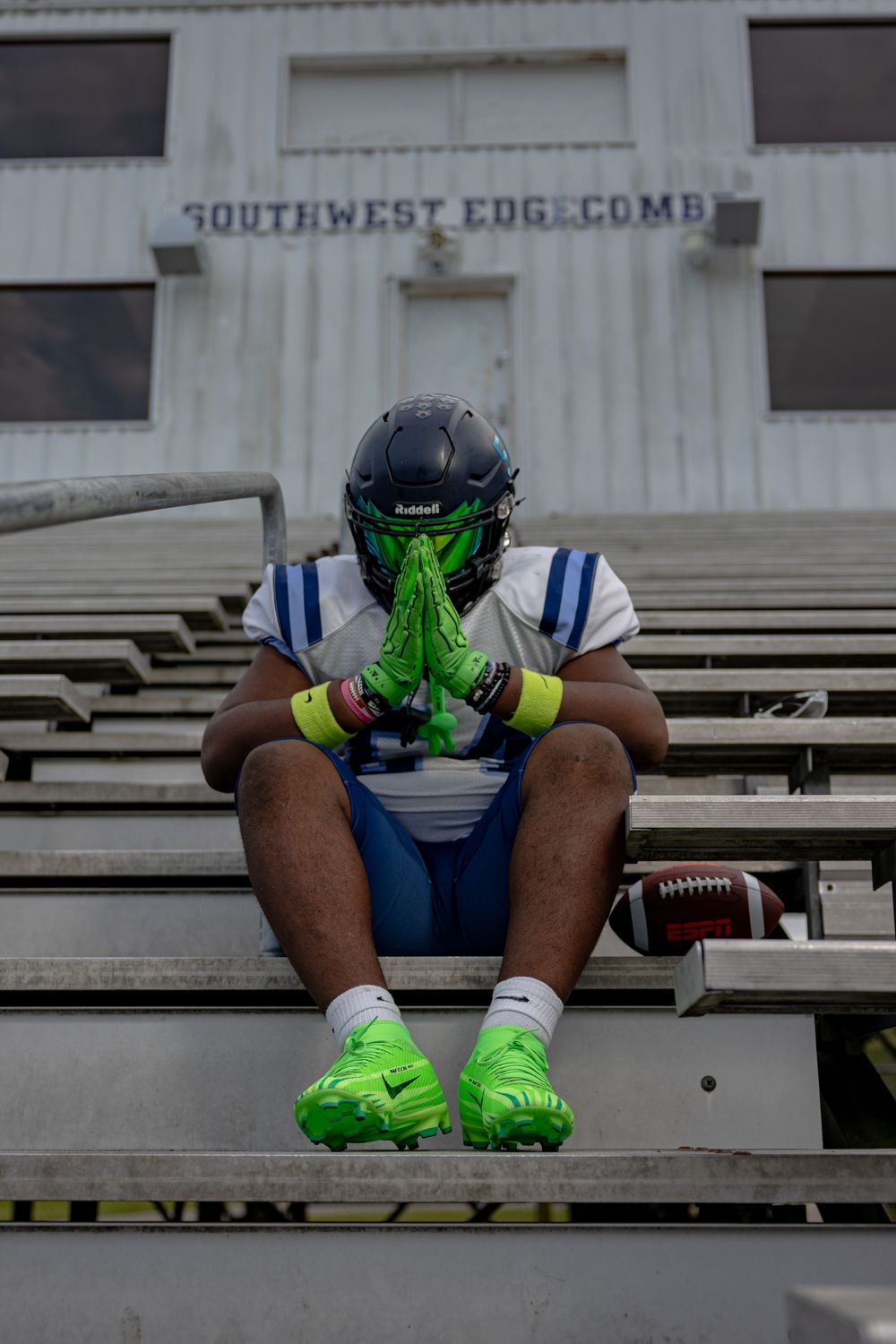 Player in football gear sitting in stadium bleachers wearing bright green accessories and navy uniform.