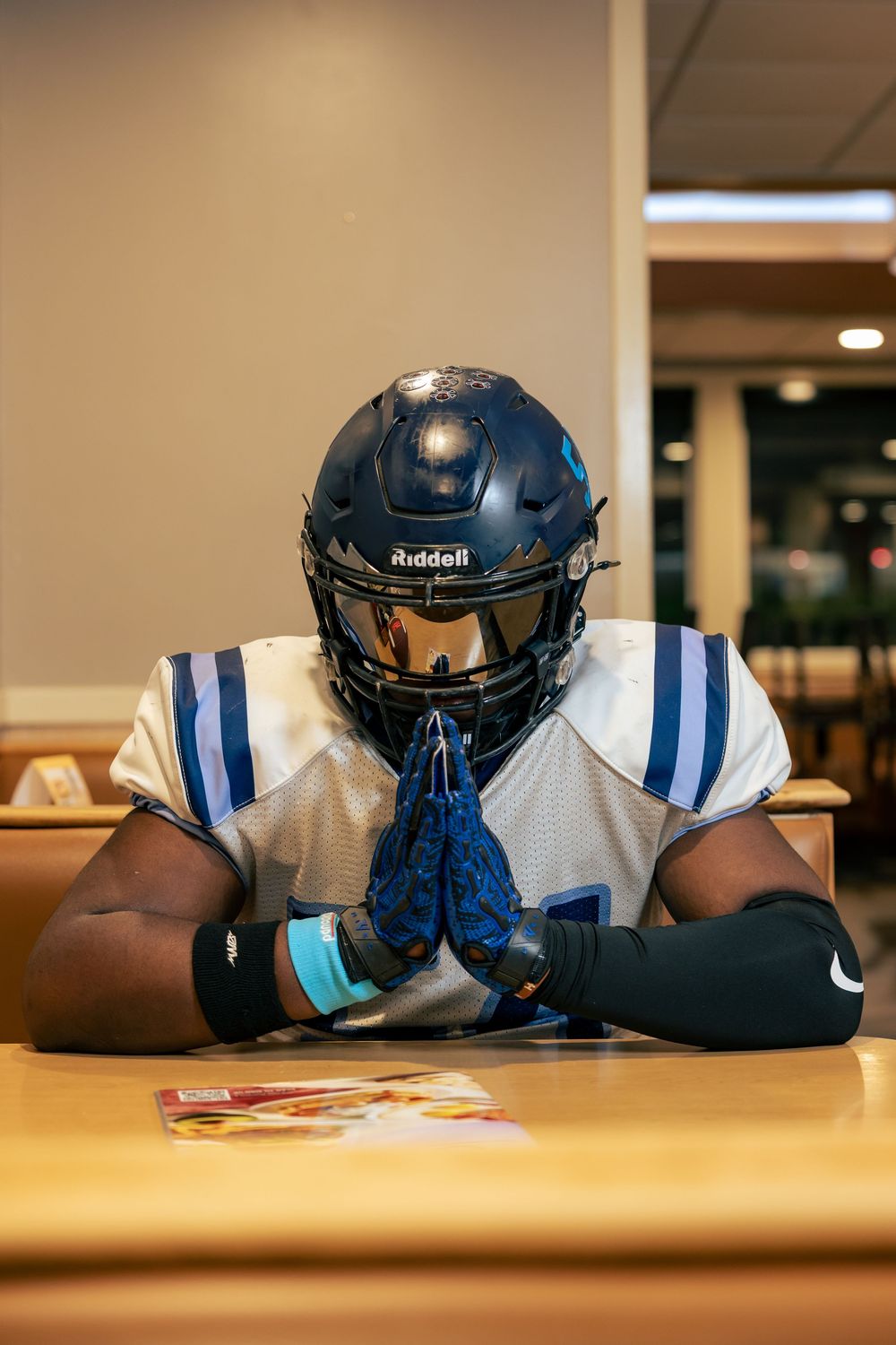 Football player in white jersey and navy helmet sitting at table with hands clasped.