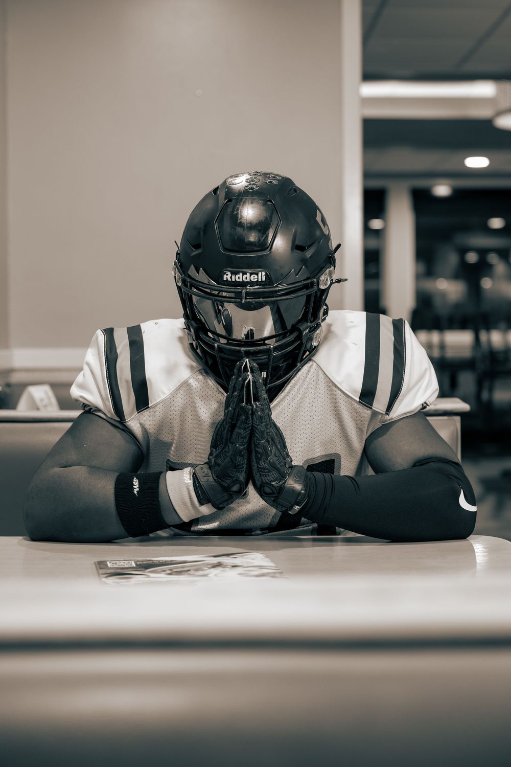 Football player wearing Riddell helmet sits at table in prayer or meditation pose.