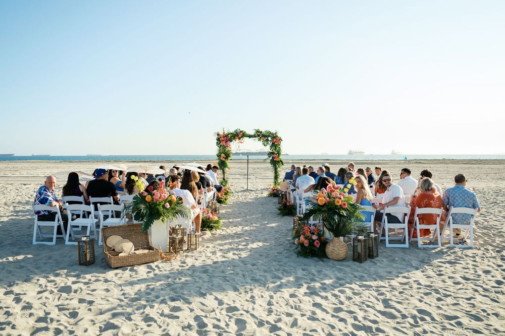Scenic beach wedding ceremony setup with white chairs and rustic decor arranged in curved rows on sandy shore.