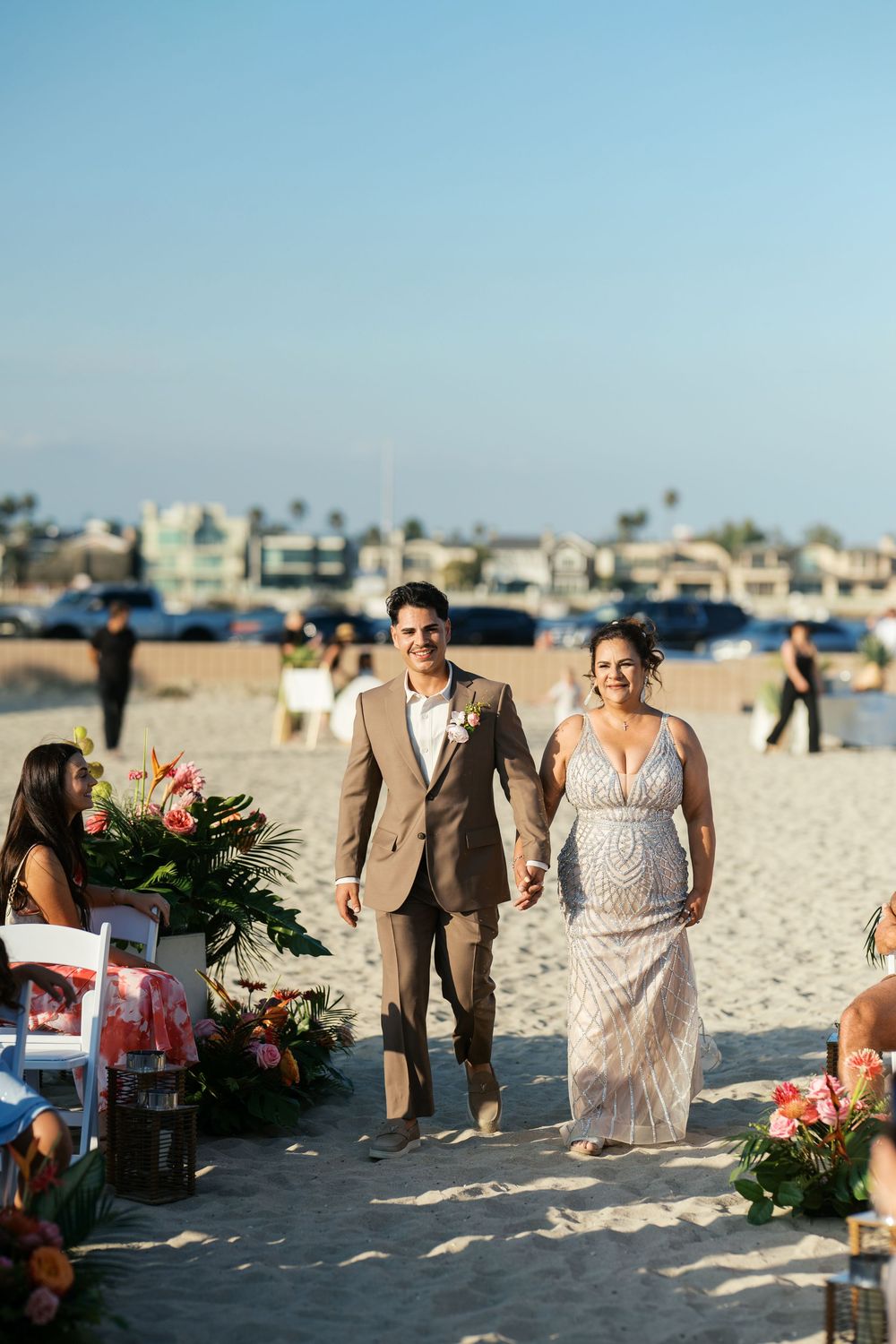 A couple walks down a sandy beach aisle adorned with tropical flowers during their oceanfront wedding ceremony.
