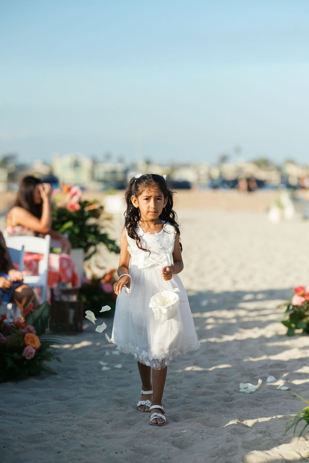 Young flower girl in white lace dress walks down beach aisle carrying flower petals at seaside wedding ceremony.