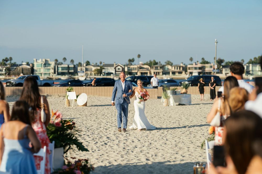 A seaside beach wedding ceremony with rows of white chairs set up on the sand as guests gather near the ocean.