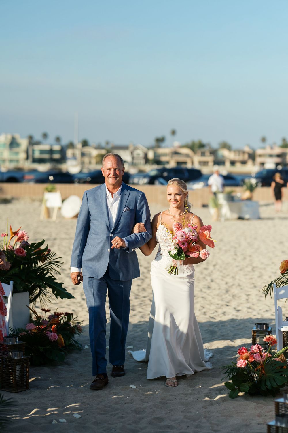 Wedding procession on sandy beach with decorated floral aisle and oceanfront houses in background.