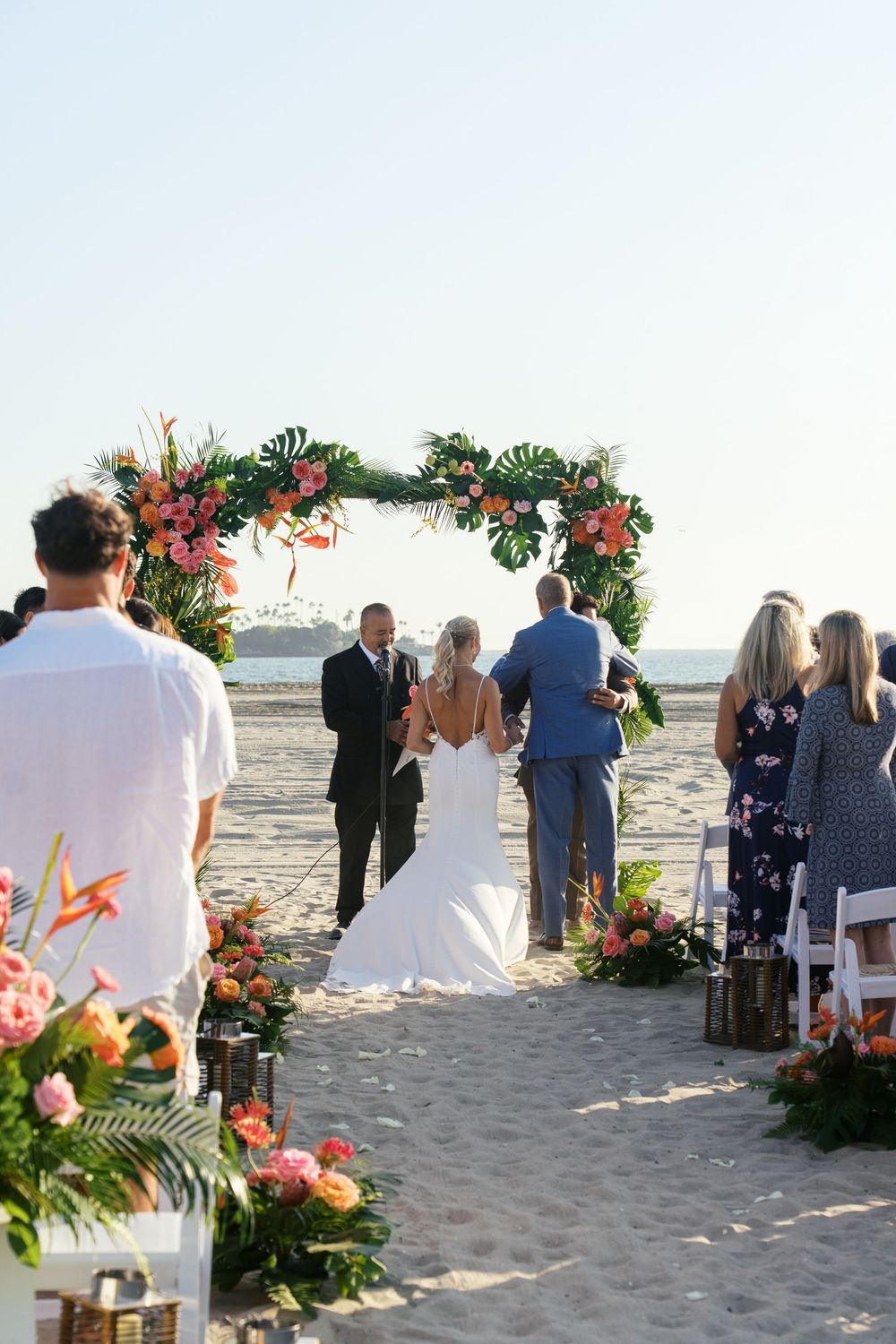 A beach wedding ceremony setup with tropical floral arrangements and guests seated on white chairs by the ocean.