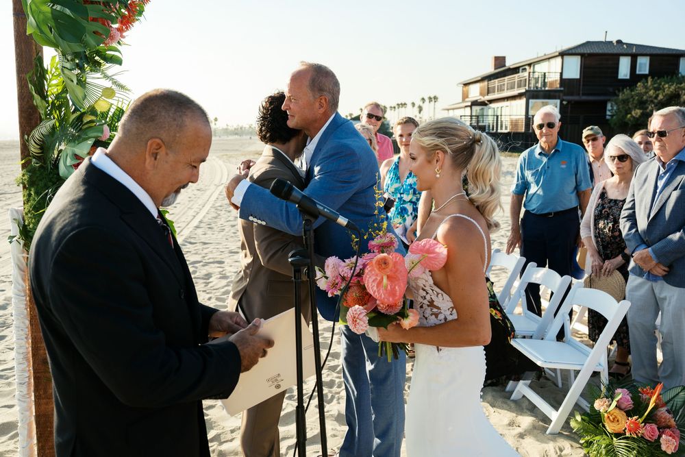 Wedding guests mingle and celebrate during an outdoor beach ceremony as the sun sets along the coast.