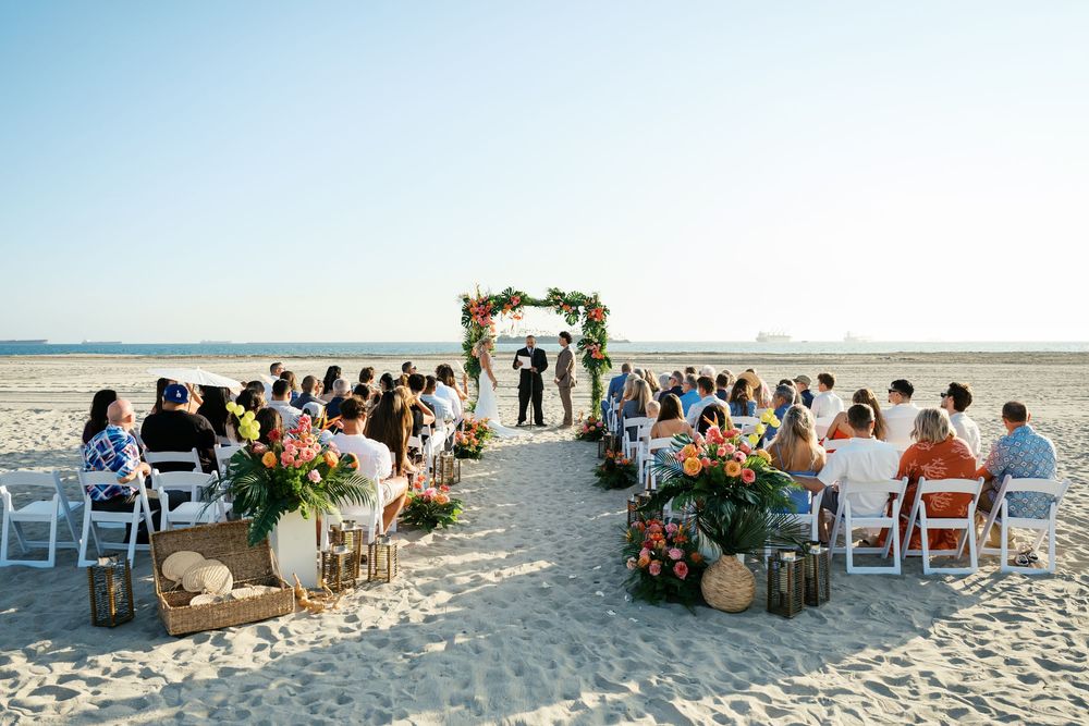 Guests seated in white chairs on a sandy beach for an oceanfront wedding ceremony at sunset.