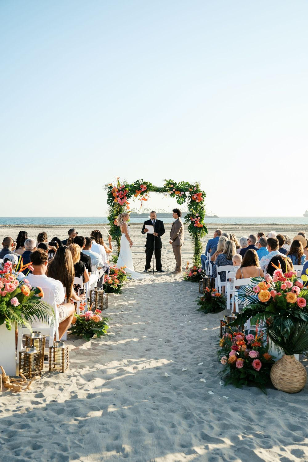 Beautiful beach wedding ceremony setup with floral arch and white chairs on sand overlooking the ocean.
